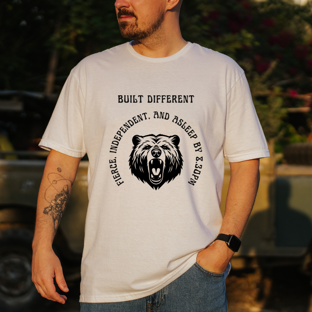 A man wearing a white graphic t-shirt with a black bear design and the text 'BUILT DIFFERENT' and 'FIERCE, INDEPENDENT, AND ASLEEP BY 8.30PM