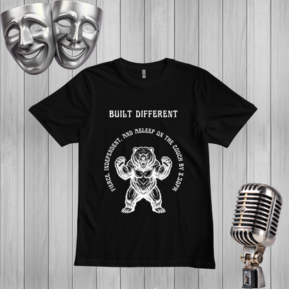 A black short-sleeve graphic T-shirt laid flat on a light gray wood plank background, with a white bear illustration and the text 'BUILT DIFFERENT' and 'Rise Independent and Asleep on the Couch by Zepan.' Silver theater masks appear in the top left, and a vintage-style microphone appears in the bottom right.