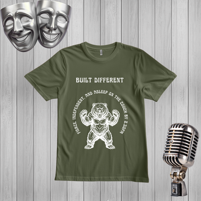 A green t-shirt with a white graphic of a bear flexing and the text 'BUILT DIFFERENT' . The shirt is displayed on a gray wood-plank background with silver comedy and theater masks in the top-left corner and a vintage silver microphone on the right.