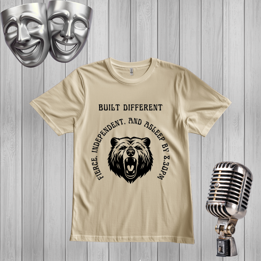 A SAND graphic t-shirt featuring a black bear illustration and the text 'BUILT DIFFERENT'The t-shirt is displayed on a gray wood plank background, styled with a silver comedy mask, a vintage microphone, and two silver theater masks in the corners.