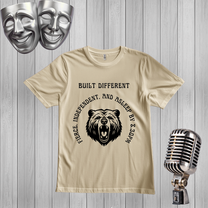 A SAND graphic t-shirt featuring a black bear illustration and the text 'BUILT DIFFERENT'The t-shirt is displayed on a gray wood plank background, styled with a silver comedy mask, a vintage microphone, and two silver theater masks in the corners.