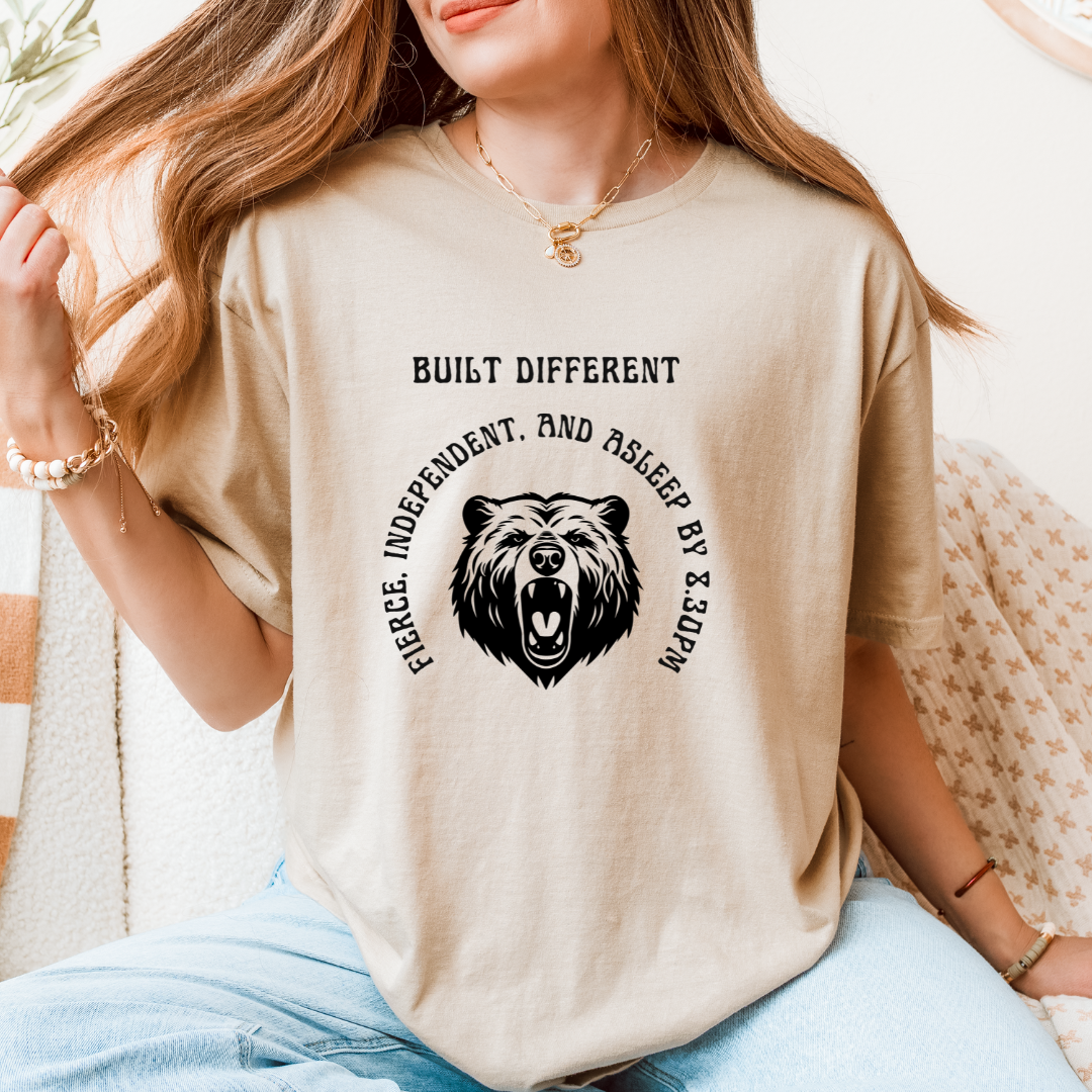 A woman wearing a SAND t-shirt with a black bear design and the text 'BUILT DIFFERENT' and 'FIERCE. INDEPENDENT.