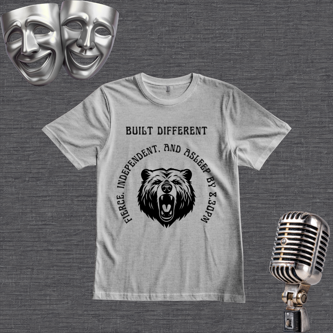 A GREY short-sleeve graphic T-shirt laid flat on a light gray wood plank background, with black text reading 'BUILT DIFFERENT' above a bear head graphic and curved text around it. Two silver theater masks appear in the top left and a vintage silver microphone appears in the bottom right.