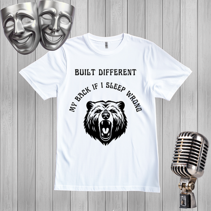 A white short-sleeve graphic t-shirt displayed on a gray wood plank background, flanked by silver theater masks on the top left and a vintage-style microphone on the bottom right. The t-shirt features a black graphic of a roaring bear with the text 'MY BACK IF I SLEEP WRONG' and 'BUILT DIFFERENT' above it.