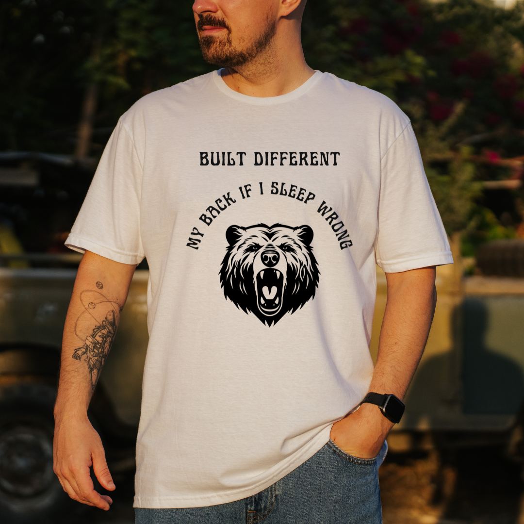 A man wearing a white graphic t-shirt with a black bear design and the text 'BUILT DIFFERENT MY BACK IF I SLEEP WRONG', photographed outdoors in warm natural light.