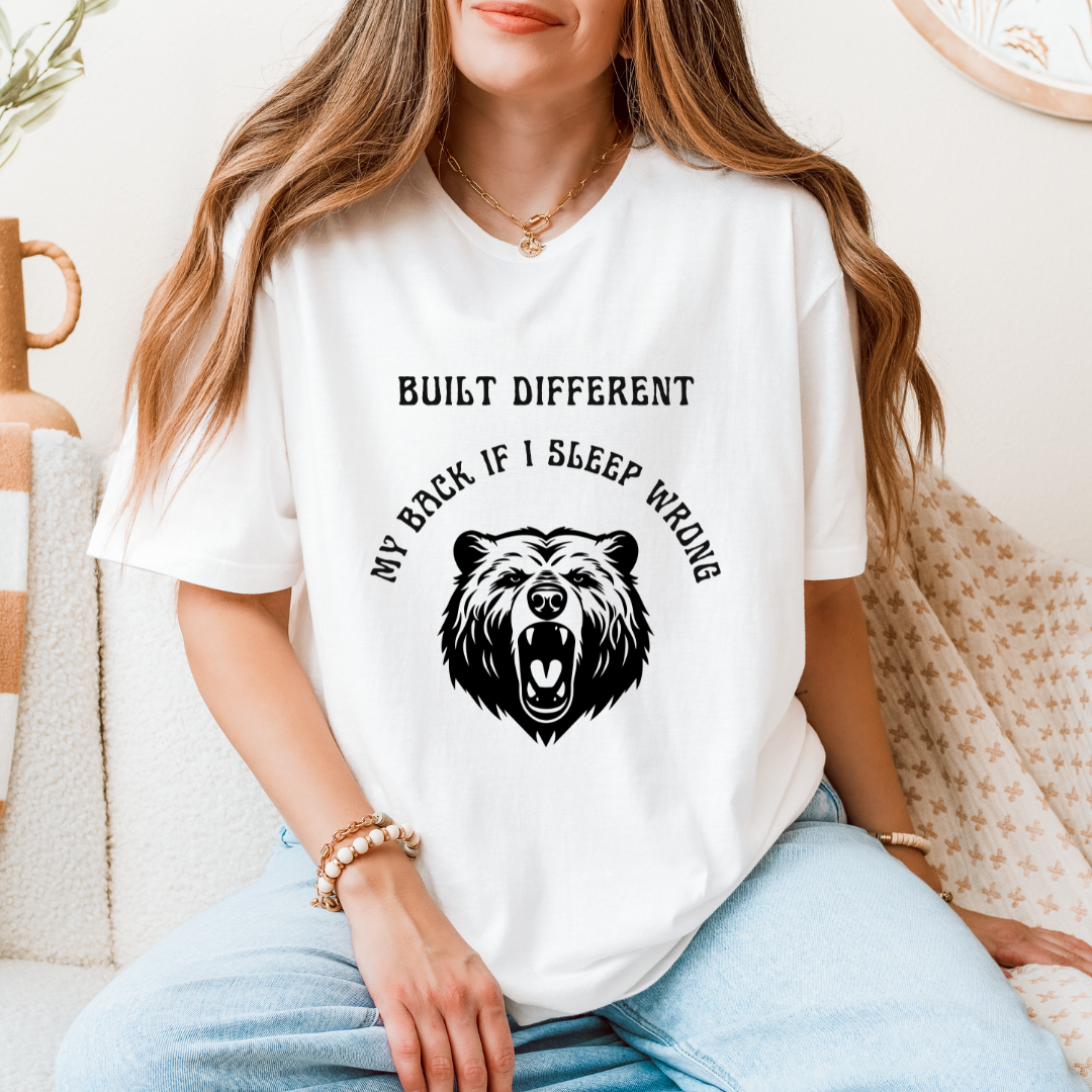A woman wearing a white graphic t-shirt with a black bear design and the text 'BUILT DIFFERENT MY BACK IF I SLEEP WRONG', paired with light blue jeans in a cozy indoor setting.