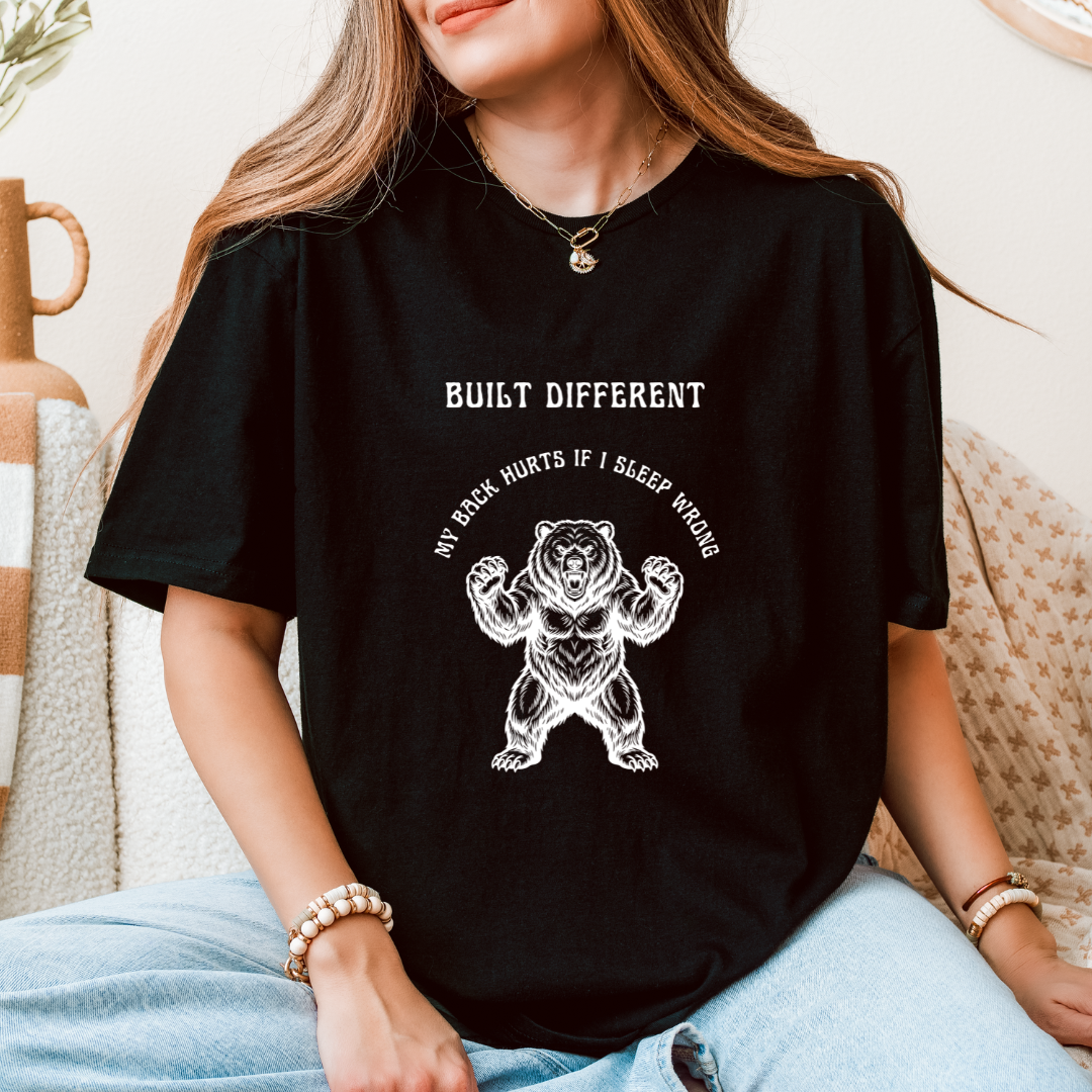 A woman wearing a black graphic t-shirt with a white bear illustration and the text 'BUILT DIFFERENT' and 'MY BACK HURTS IF I SLEEP WRONG'.