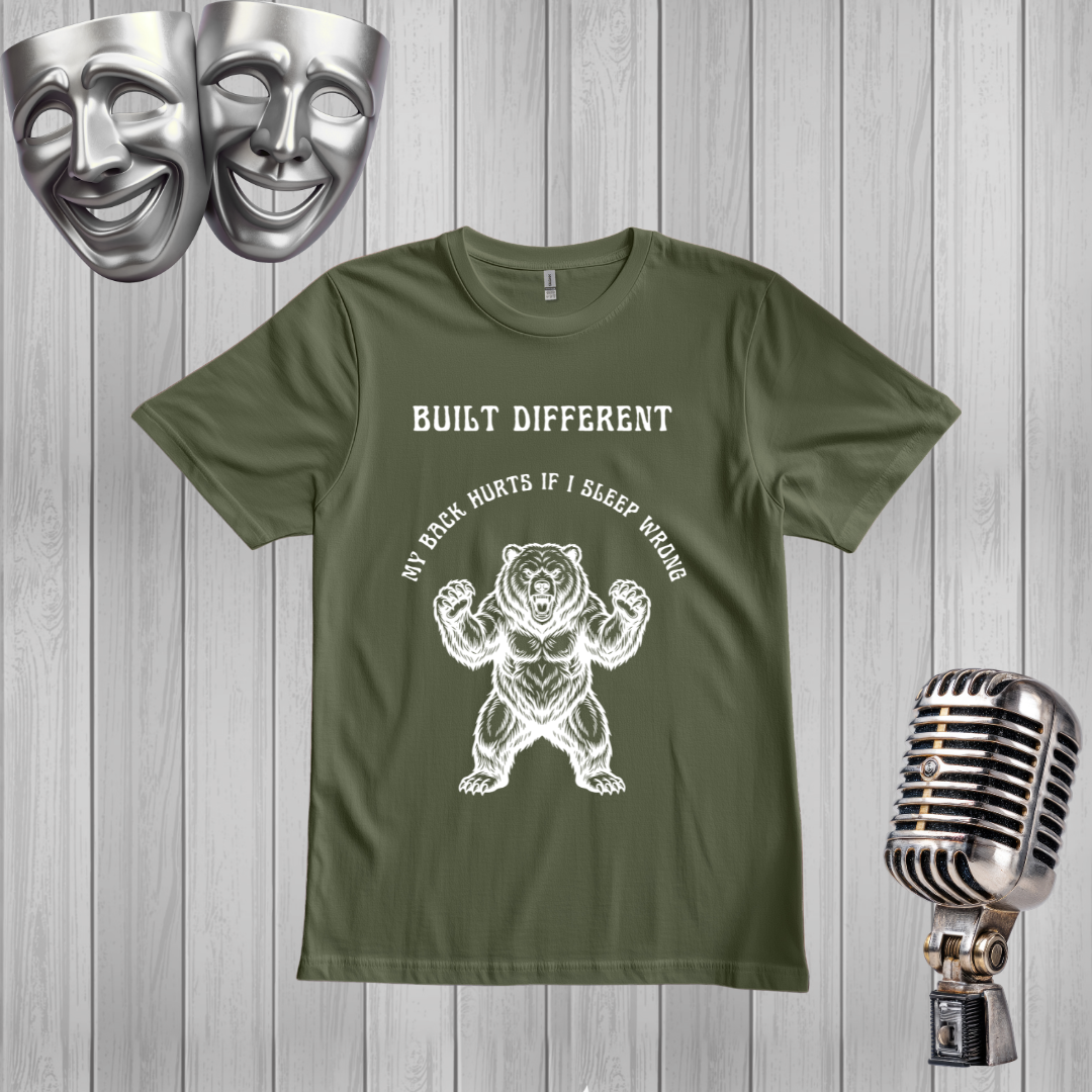 A Green short-sleeve graphic t-shirt displayed on a gray wood plank background, flanked by silver theater masks on the top left and a vintage-style microphone on the bottom right. The t-shirt features a black graphic of a roaring bear with the text 'MY BACK IF I SLEEP WRONG' and 'BUILT DIFFERENT' above it.