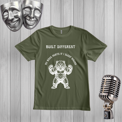 A Green short-sleeve graphic t-shirt displayed on a gray wood plank background, flanked by silver theater masks on the top left and a vintage-style microphone on the bottom right. The t-shirt features a black graphic of a roaring bear with the text 'MY BACK IF I SLEEP WRONG' and 'BUILT DIFFERENT' above it.