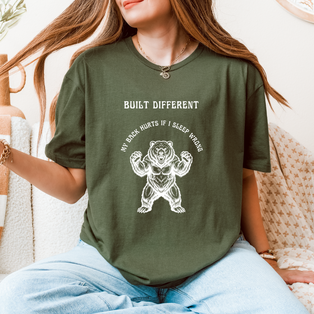 A woman wearing a green graphic t-shirt with a white bear illustration and the text 'BUILT DIFFERENT' and 'MY BACK HURTS IF I SLEEP WRONG'.
