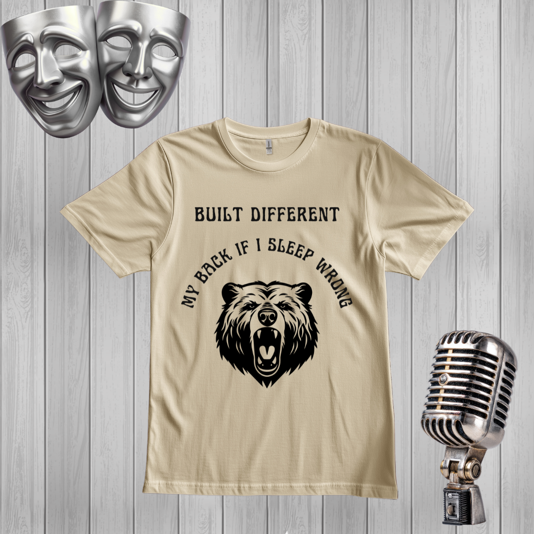 A sand graphic t-shirt featuring a black bear illustration and the text 'BUILT DIFFERENT MY BACK IF I SLEEP WRONG', displayed on a gray wood plank background with silver comedy and tragedy masks and a vintage microphone as props.