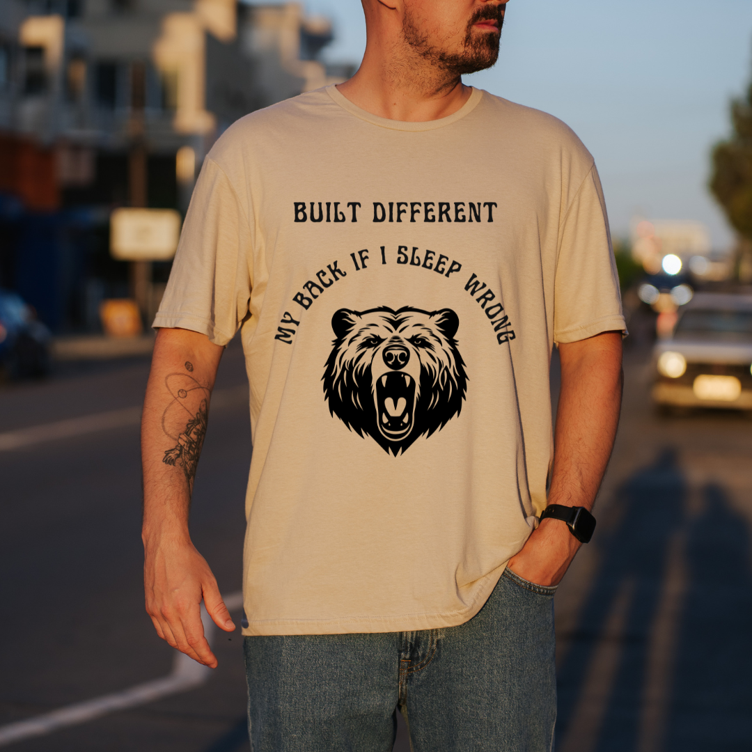 A man wearing a sand graphic t-shirt with a black bear design and the text 'BUILT DIFFERENT MY BACK IF I SLEEP WRONG', standing on a blurred street at sunset.
