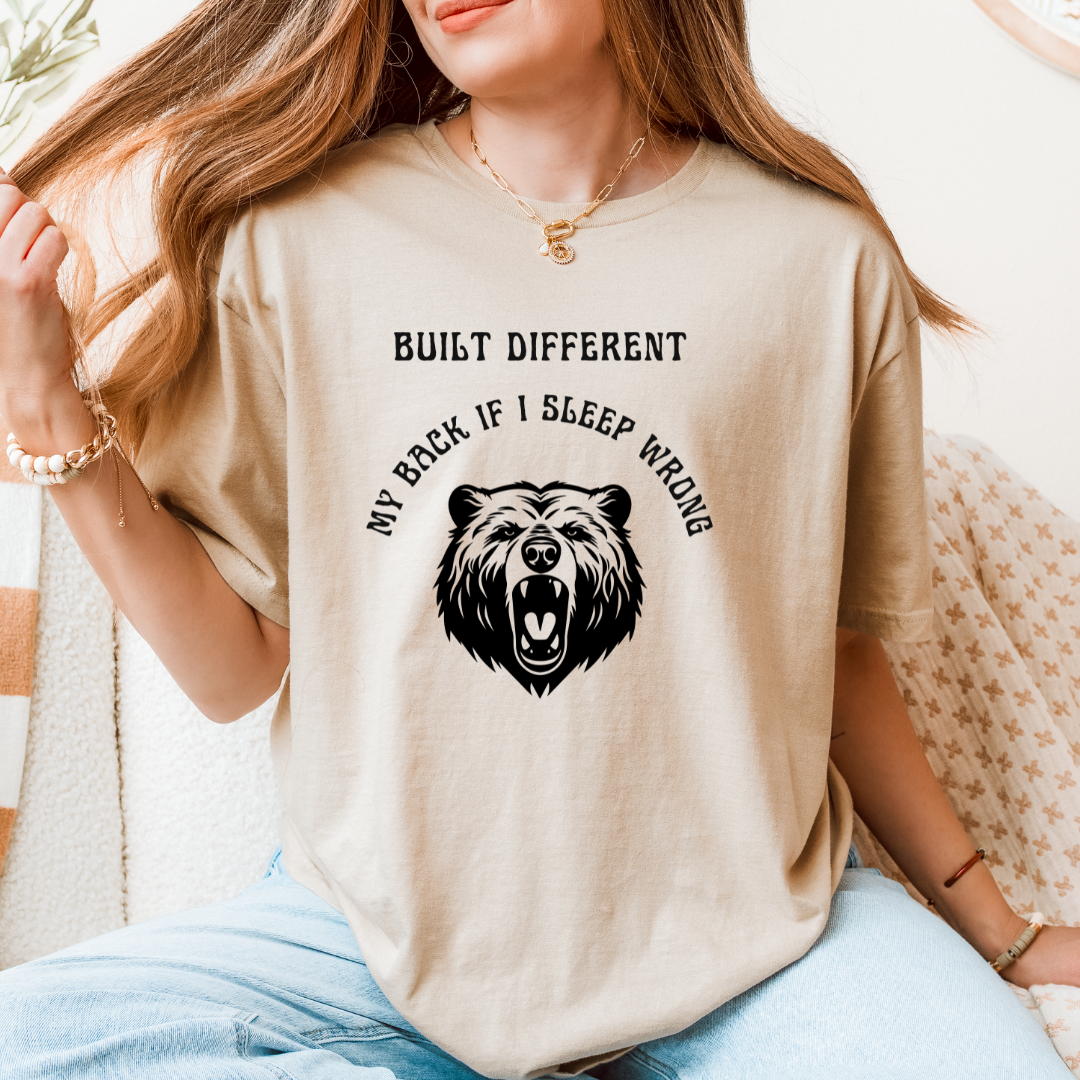 A woman wearing a sand graphic t-shirt with a black bear design and the text 'BUILT DIFFERENT MY BACK IF I SLEEP WRONG'.
