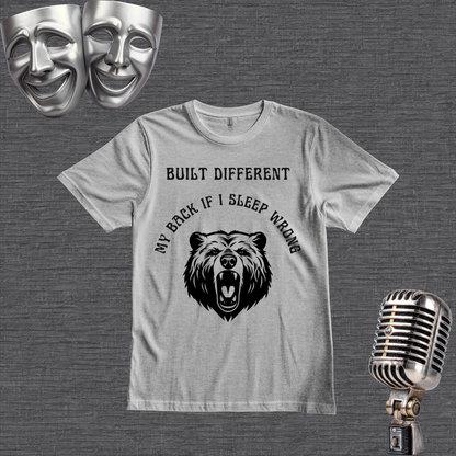 A grey short-sleeve graphic t-shirt displayed on a gray wood plank background, flanked by silver theater masks on the top left and a vintage-style microphone on the bottom right. The t-shirt features a black graphic of a roaring bear with the text 'MY BACK IF I SLEEP WRONG' and 'BUILT DIFFERENT' above it.