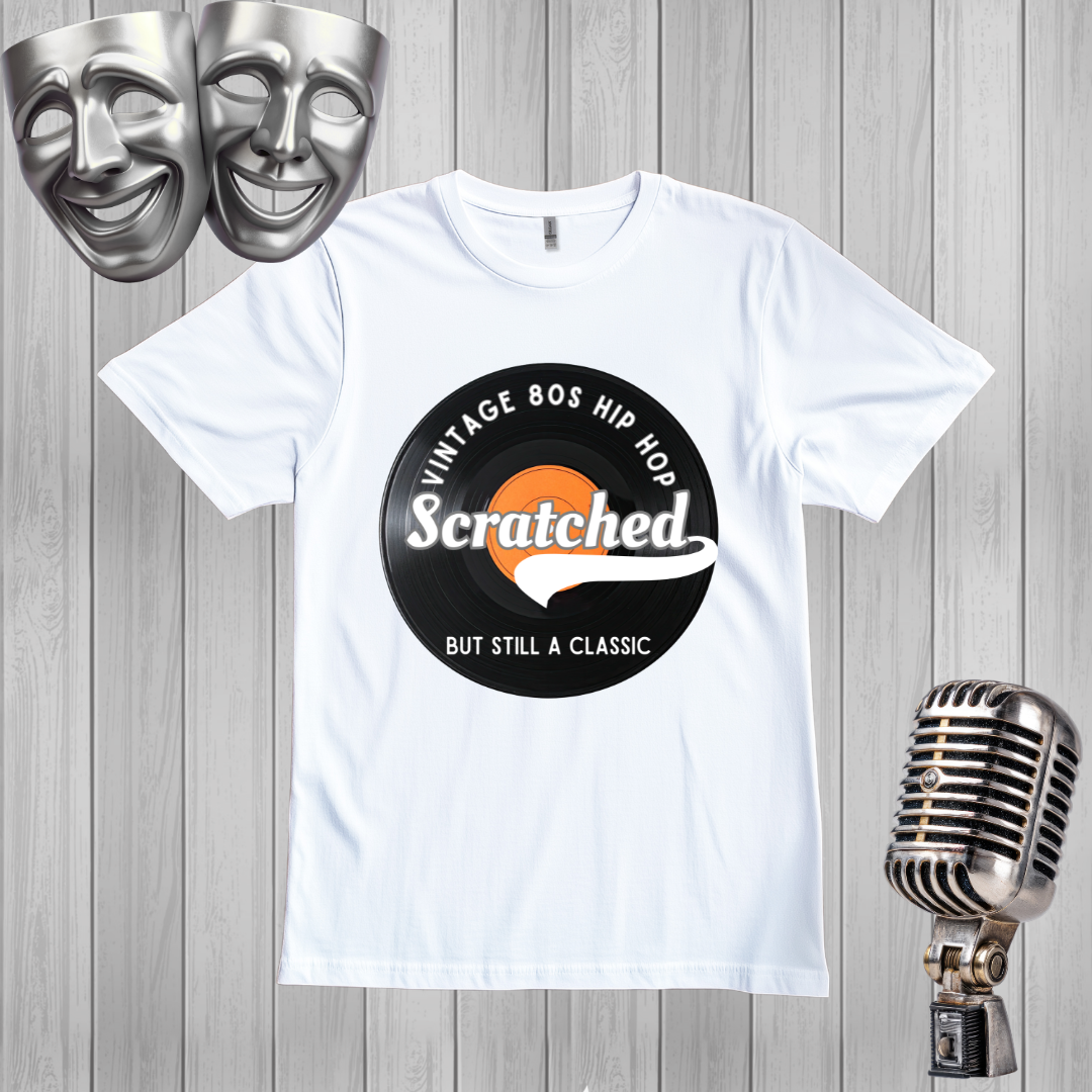 A white short-sleeve graphic T-shirt laid flat on a gray wood plank background, featuring a circular black-and-orange print that reads 'Vintage 80s Hip Hop' and 'Scratched But Still A Classic.' Silver comedy and microphone props appear around the shirt.