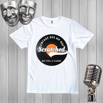 A white short-sleeve graphic T-shirt laid flat on a gray wood plank background, featuring a circular black-and-orange print that reads 'Vintage 80s Hip Hop' and 'Scratched But Still A Classic.' Silver comedy and microphone props appear around the shirt.