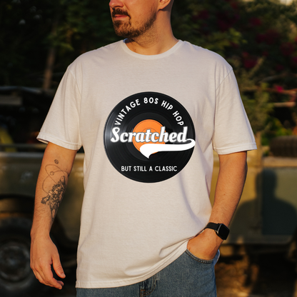 A man wearing a white graphic t-shirt with a black circular 'Vintage 80s Hip Hop' design featuring the word 'Scratched' in white over an orange circle, paired with blue jeans in an outdoor setting.