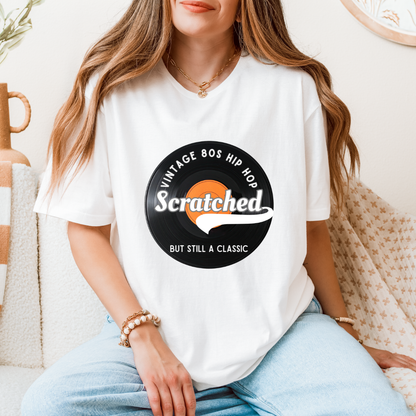 A woman wearing a white graphic t-shirt with a black and orange circular 'Vintage 80s Hip Hop' design featuring the word 'Scratched'. She is sitting on a cozy beige couch with a patterned throw blanket.
