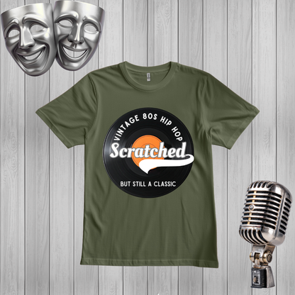 A green crewneck t-shirt featuring a circular graphic print that reads 'VINTAGE 80S HIP HOP Scratched BUT STILL A CLASSIC' over a black vinyl record and orange egg graphic. The shirt is displayed on a gray wood plank background, flanked by silver comedy and tragedy masks on the top left and a vintage silver microphone on the bottom right.