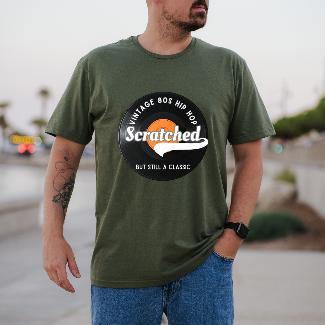 A man wearing a green t-shirt with a circular graphic that reads 'VINTAGE 80S HIP HOP Scratched BUT STILL A CLASSIC' in white and orange, paired with blue jeans, standing outdoors with a blurred background.
