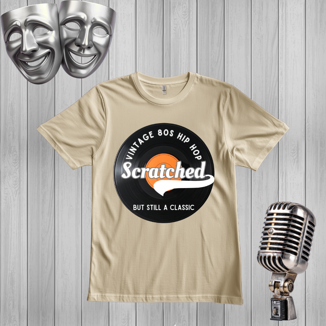 A sand graphic t-shirt featuring a retro 80s hip hop design with the word 'Scratched' on a black circular graphic, displayed on a gray wood plank background with silver comedy masks and a vintage microphone.