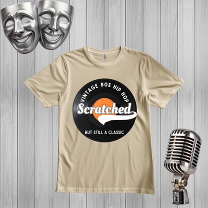 A sand graphic t-shirt featuring a retro 80s hip hop design with the word 'Scratched' on a black circular graphic, displayed on a gray wood plank background with silver comedy masks and a vintage microphone.