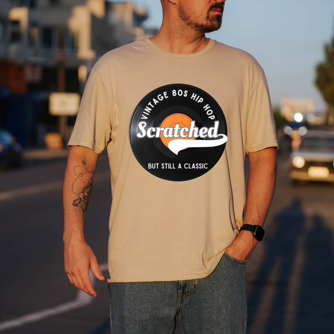A man wearing a sand graphic t-shirt with a retro 80s hip-hop vinyl record design that reads 'Scratched BUT STILL A CLASSIC'. The background is a blurred street scene at sunset.