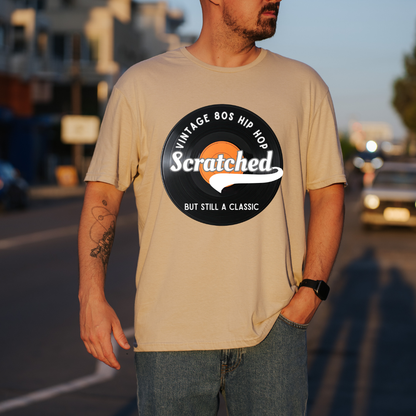 A man wearing a sand graphic t-shirt with a retro 80s hip-hop vinyl record design that reads 'Scratched BUT STILL A CLASSIC'. The background is a blurred street scene at sunset.