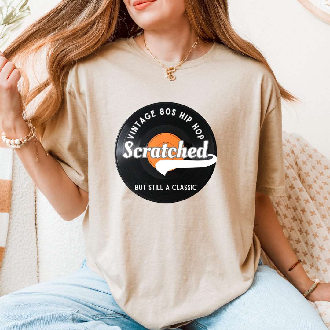 A woman wearing a sand graphic t-shirt with a black circular 'Vintage 80s Hip Hop' design featuring the word 'Scratched' in white over an orange circle, paired with light blue jeans.
