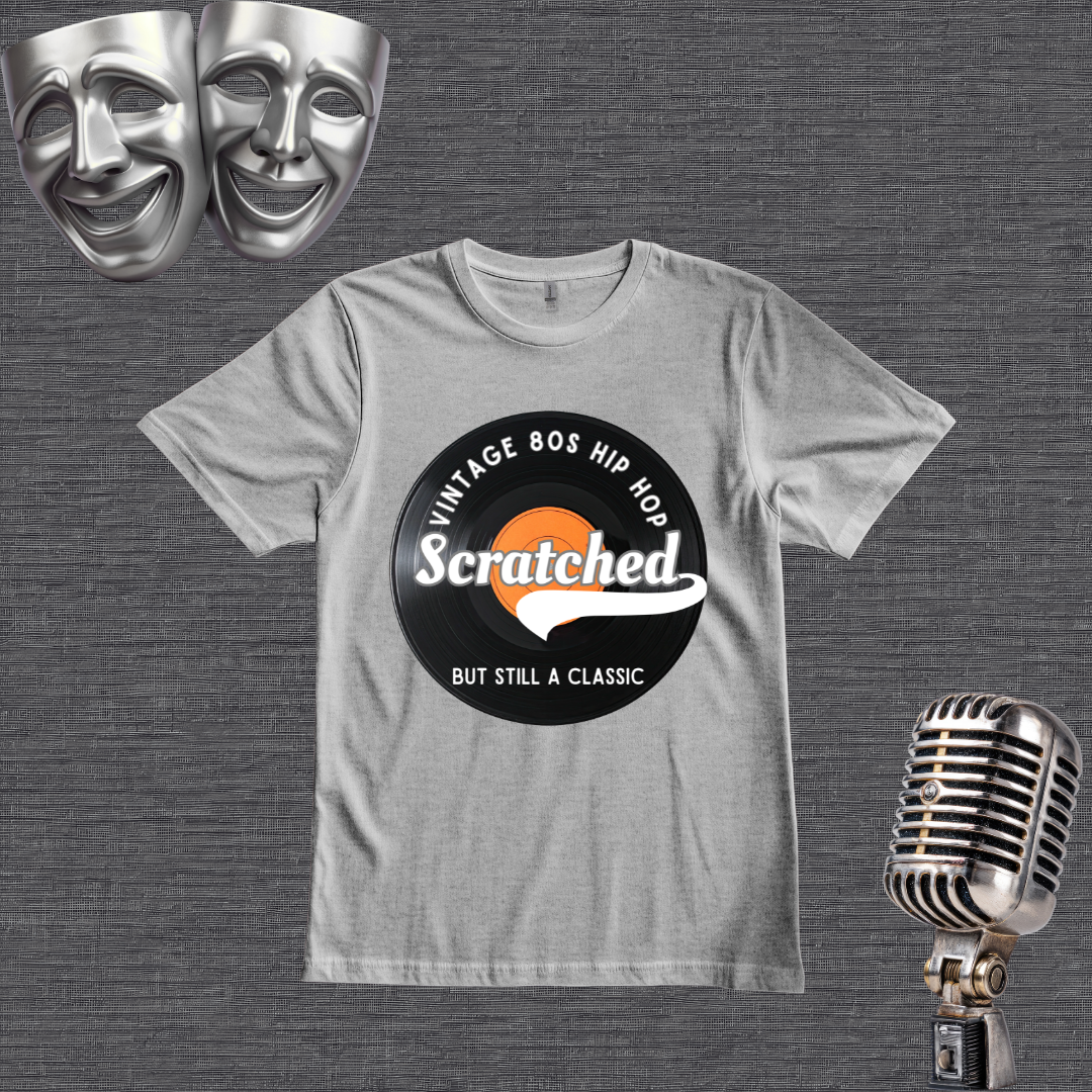 A light gray crewneck t-shirt featuring a retro 1980s hip-hop graphic with the word 'Scratched' on a vinyl record design. The shirt is displayed on a textured gray background with silver comedy and tragedy masks in the top-left corner and a vintage silver microphone in the bottom-right corner.