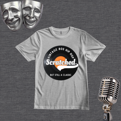 A light gray crewneck t-shirt featuring a retro 1980s hip-hop graphic with the word 'Scratched' on a vinyl record design. The shirt is displayed on a textured gray background with silver comedy and tragedy masks in the top-left corner and a vintage silver microphone in the bottom-right corner.