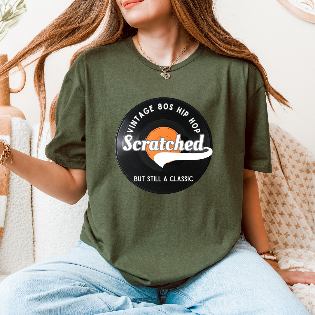 A woman wearing an olive green graphic t-shirt with a retro vinyl record design that reads 'VINTAGE BOS HIP Hop Scratched BUT STILL A CLASSIC'.