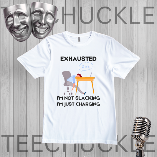 A white graphic t-shirt displayed on a gray wood plank background, featuring a cartoon illustration of a person sleeping at a desk with the text 'EXHAUSTED I'M NOT SLACKING I'M JUST CHARGING'. The image includes decorative props like silver theater masks and a vintage microphone, 