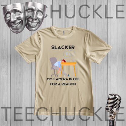 A sand graphic t-shirt displayed on a gray wood plank background, featuring a cartoon illustration of a person sleeping at a desk with the text 'SLACKER, MY CAMERA IS OFF FOR A REASON'. The image includes decorative props like silver theater masks and a vintage microphone, 