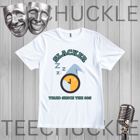A white short-sleeve graphic t-shirt displayed on a gray wood-plank background, featuring a 90s-themed design with the word 'SLACKER', 'Z Z Z' sleep marks, a clock graphic, and 'TIRED SINCE THE 90S'. The image includes decorative props like silver theater masks and a vintage microphone