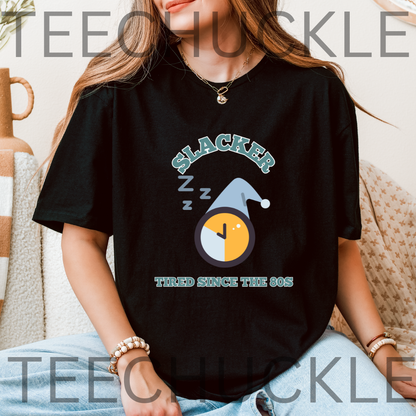 A woman wearing a black graphic t-shirt with a 'Slacker' sleeping clock design, sitting in a cozy indoor setting. The t-shirt features a graphic of a clock with a hat and 'Z' sleep sounds, with the text 'SLACKER TIRED SINCE THE 80S'.