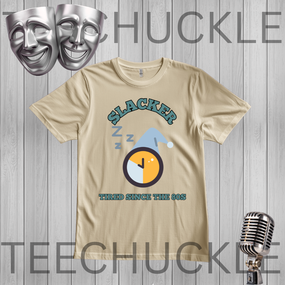 A sand short-sleeve graphic t-shirt displayed on a gray wood-plank background, featuring a 90s-themed design with the word 'SLACKER', 'Z Z Z' sleep marks, a clock graphic, and 'TIRED SINCE THE 80S'. The image includes decorative props like silver theater masks and a vintage microphone