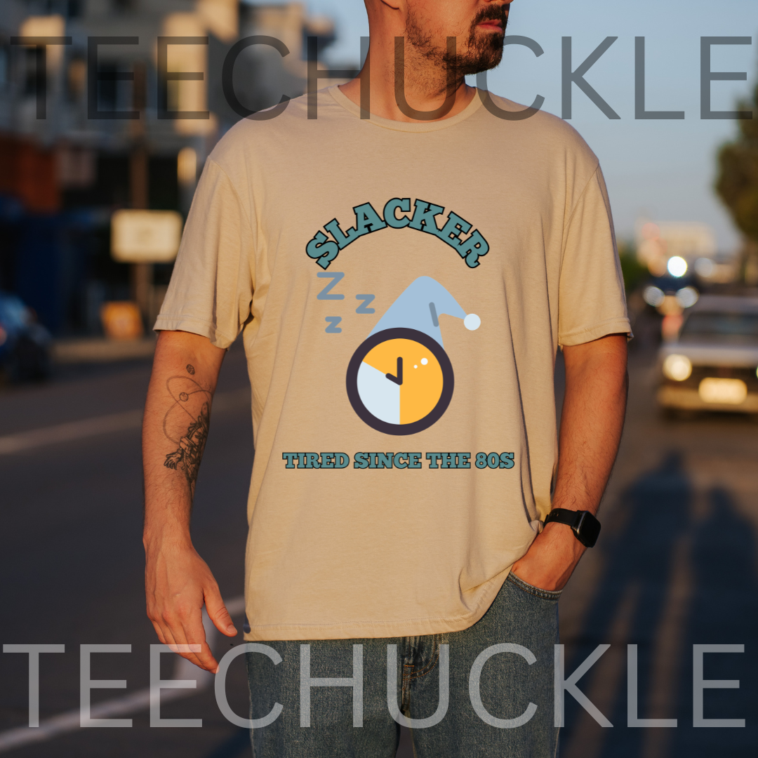A man wearing a sand graphic t-shirt with a 'Slacker' sleeping clock design, standing outdoors on a street at sunset. 