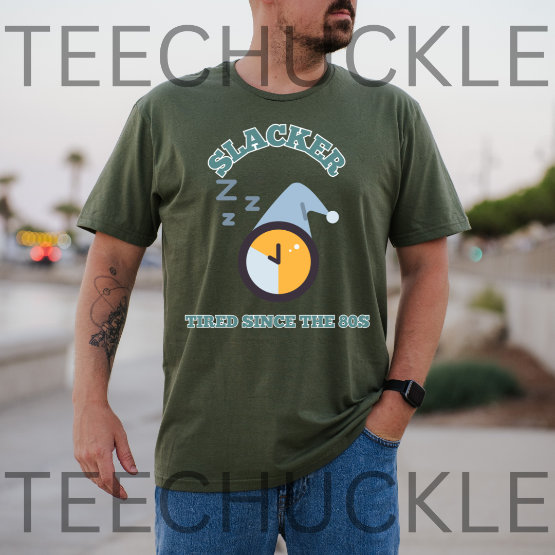 A man wearing a green graphic t-shirt with a 'Slacker Tired Since The 80s' design, standing outdoors with a blurred background. The t-shirt features a clock graphic with a sleeping hat and 'Z' marks.