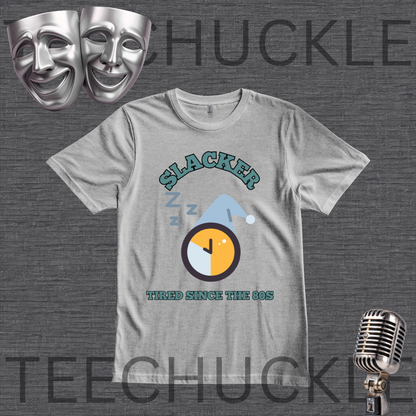 A gray graphic t-shirt featuring a 'Slacker' theme with a sleeping clock and the text 'Tired Since the 80s'. The t-shirt is displayed on a dark gray textured background with theatrical comedy masks, a vintage microphone