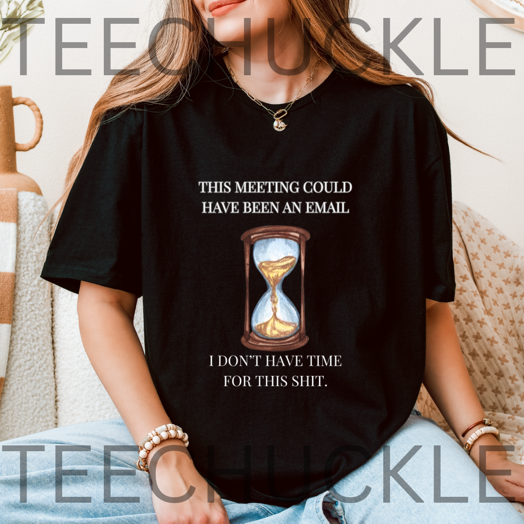 A woman wearing a black graphic t-shirt with the text 'THIS MEETING COULD HAVE BEEN AN EMAIL' above an hourglass illustration and 'I DON'T HAVE TIME FOR THIS SHIT.' below, set against a cozy indoor background with a beige sofa 
