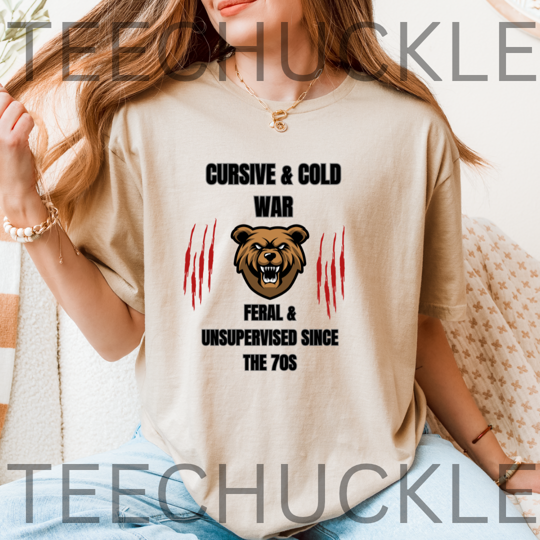 A woman wearing a beige graphic t-shirt with a brown bear design and text reading 'CURSIVE & COLD WAR FERAL & UNSUPERVISED SINCE THE 70S'. 