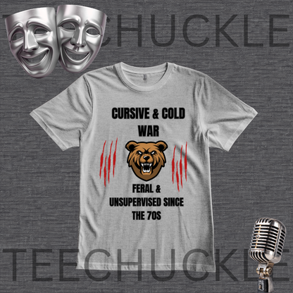 A gray graphic t-shirt displayed on a dark gray textured background, featuring a bear graphic and the text 'CURSIVE & COLD WAR FERAL & UNSUPERVISED SINCE THE 70S'. The image includes decorative props like silver theater masks, a vintage microphone