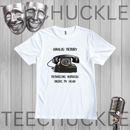 A white short-sleeve graphic T-shirt laid flat on a gray wood-plank background. The shirt has a black printed design featuring a vintage rotary telephone and the text 'ANALOGIC MEMORY' and 'MEMORIZING NUMBERS INSIDE MY HEAD.