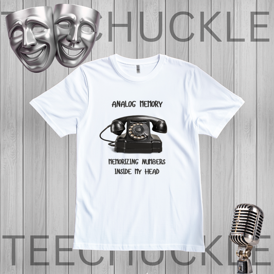 A white short-sleeve graphic T-shirt laid flat on a gray wood-plank background. The shirt has a black printed design featuring a vintage rotary telephone and the text 'ANALOGIC MEMORY' and 'MEMORIZING NUMBERS INSIDE MY HEAD.