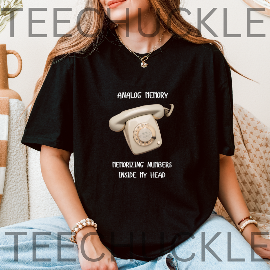 A woman wearing a black graphic t-shirt with a retro rotary phone design and the text 'ANALOGUE MEMORY' and 'MEMORIZING NUMBERS INSIDE MY HEAD'.