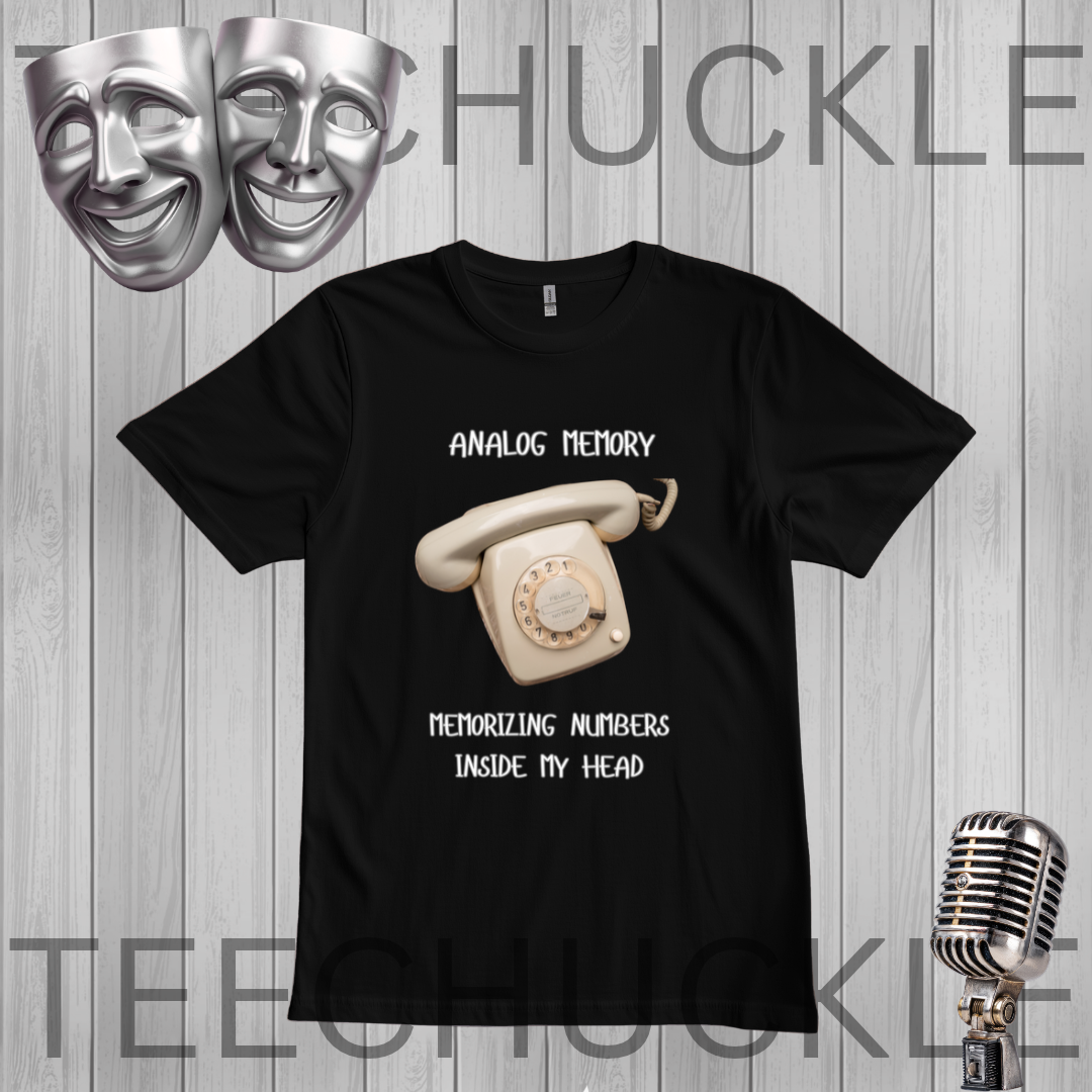 A black short-sleeve graphic t-shirt featuring a vintage cream rotary phone and the text 'ANALOGIC MEMORY' and 'MEMORIZING NUMBERS INSIDE MY HEAD'. The shirt is displayed on a gray wood plank background with a silver theater mask, a vintage microphone,