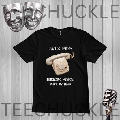 A black short-sleeve graphic t-shirt featuring a vintage cream rotary phone and the text 'ANALOGIC MEMORY' and 'MEMORIZING NUMBERS INSIDE MY HEAD'. The shirt is displayed on a gray wood plank background with a silver theater mask, a vintage microphone,