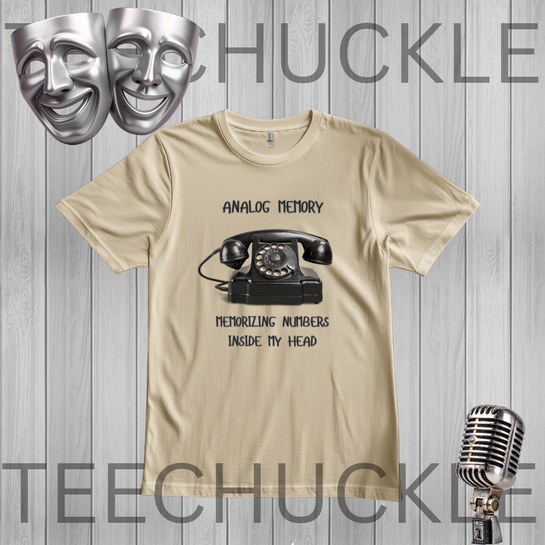 A sand graphic t-shirt featuring a black retro rotary telephone and the text 'ANALOGIC MEMORY' and 'MEMORIZING NUMBERS INSIDE MY HEAD'. The t-shirt is displayed on a gray wood-plank background with decorative silver theater masks and a vintage microphone,