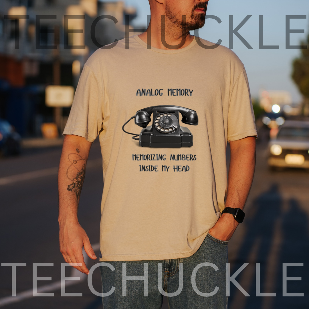 A man wearing a sand graphic t-shirt with a black rotary telephone design and the text 'ANALOGUE MEMORY MEMORIZING NUMBERS INSIDE MY HEAD'. The photo is taken outdoors on a street at sunset, 